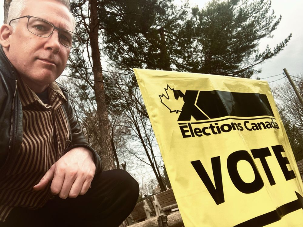 A bespectacled man kneels by an Elections Canada sign at an advance polling station.