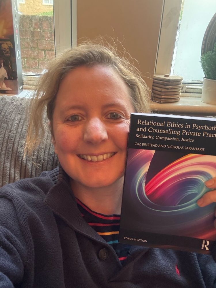 Picture of Caz wearing a striped multi-coloured top and blue jumper holding the book as described in the text - ‘Relational Ethics in Psychotherapy and Counselling Private Practice: Solidarity, Compassion, Justice’. We can also see in the background coasters, a book on magic and a mini Xmas tree 