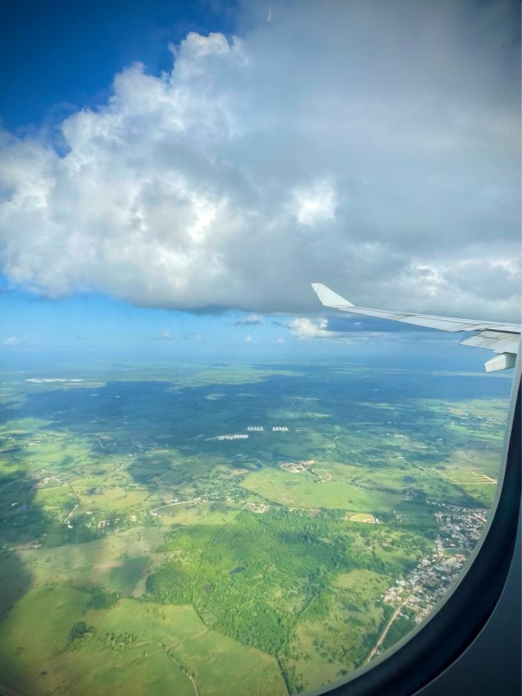 Vue au travers d’un hublot d’avion, une aile d’avion, un ciel avec quelques nuages, et la République dominicaine 🇩🇴 au-dessous