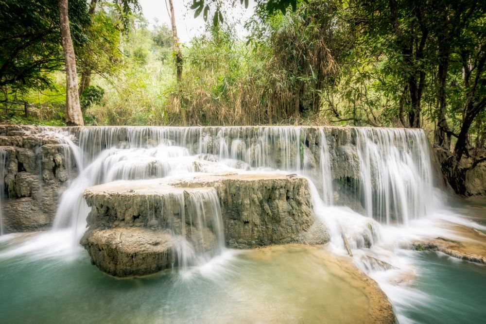 Colour photograph showing a small set of waterfalls. The water is cascading over several rocks. The background is a treed jungle area. 