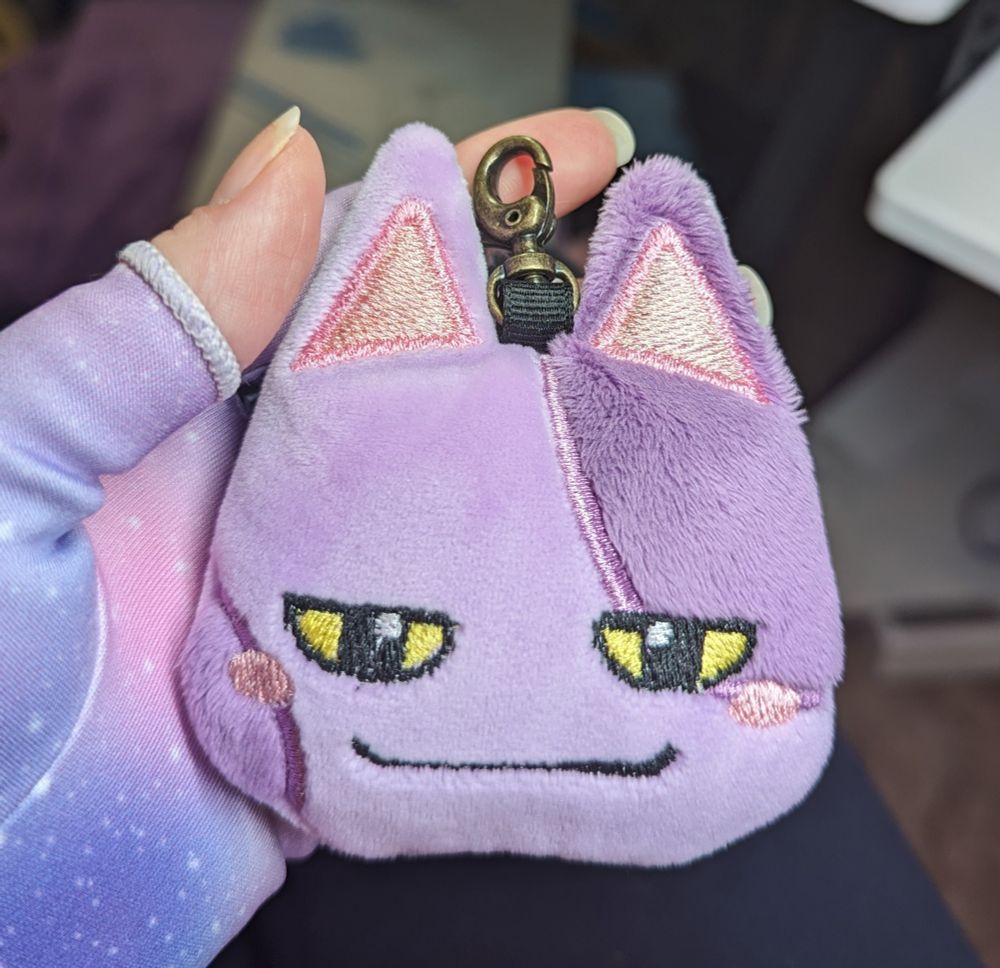 A plush keychain of the purple and lavender cat Bob from the Animal Crossing video game series.  Bob has yellow and black eyes and a smirk embroidered in black thread. The keychain is being held in palm of a white person's left hand wearing a lavender pastel galaxy print compression glove.