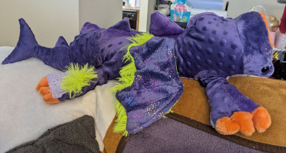 Image 2 A purple plush creature with four purple legs with orange claws, green wings and a large orange mouth. It is stretched out horizontally on the back of a sofa.
