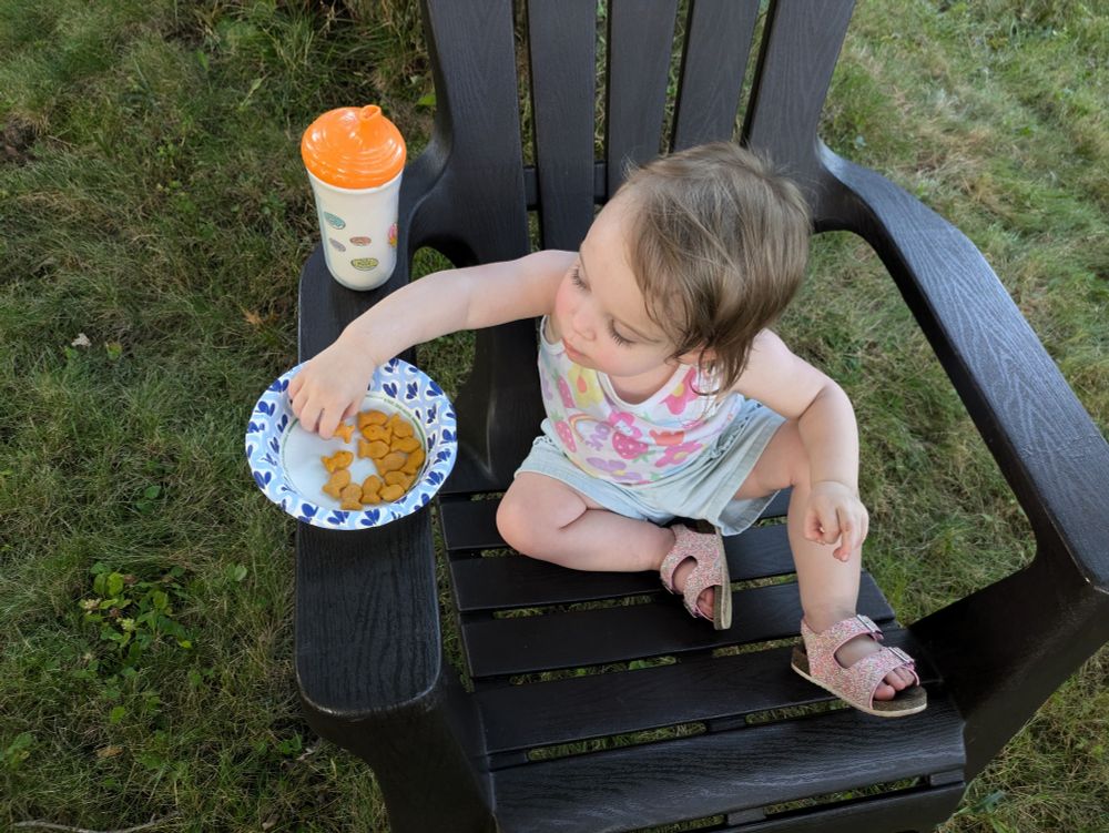 The Grem (AKA Victoria) sitting in an oversized chair outside, eating goldfish crackers, and sipping juice from her Garfield cup