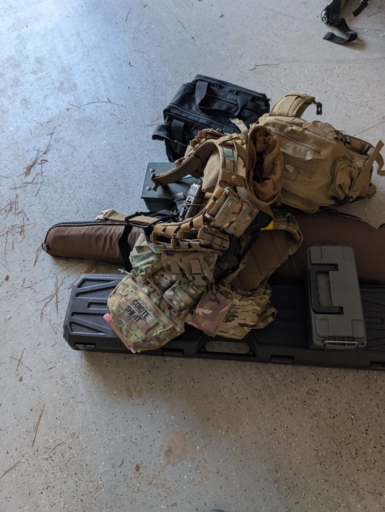 A pile of equipment and gun bags, the patches on the plate carrier read "Coyote" and "She/It"