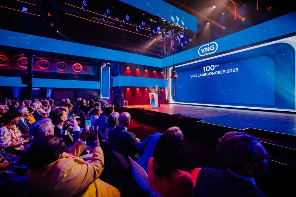 Deelnemers aan het VNG congres kijken naar de spreker op het podium. Er hangt een bel in het midden.