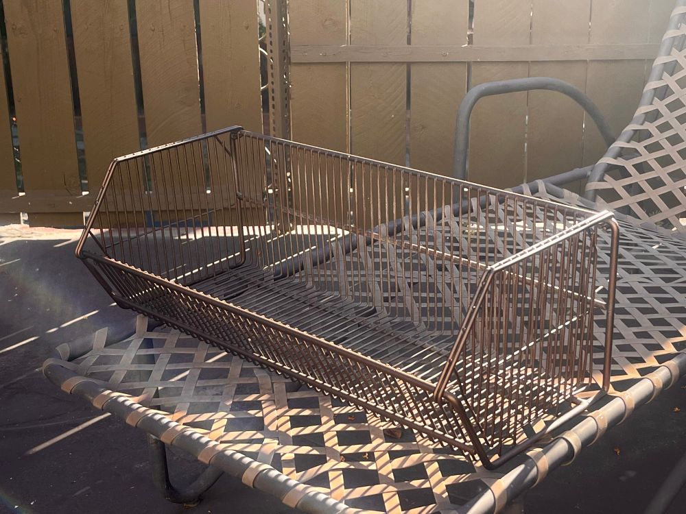A large metal wire basket from a retail craft store, about two feet wide and likely used for holding bundles of yarn, sitting on a deck chair in the sunlight. It’s been spray painted a rustic bronze.