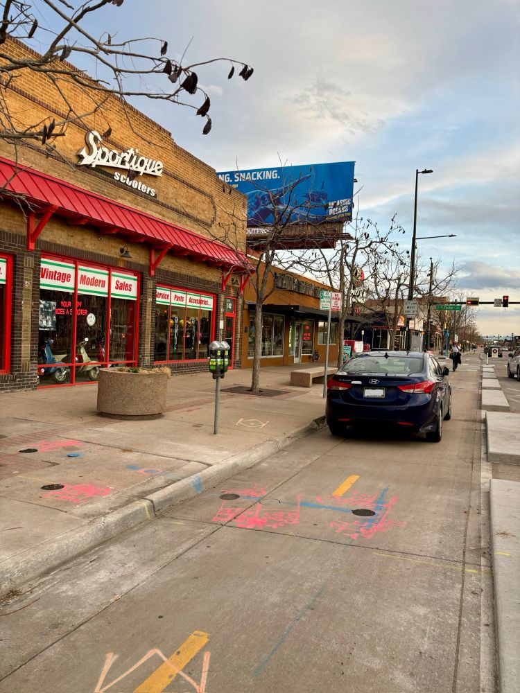 Photo of a blue car parked in the middle of a two-way protected bike lane. The sidewalk is on the left and to the right are concrete blocks/curbs that separate the street from the bike lane. Designated parking is to the right of these blocks and there was plenty of space