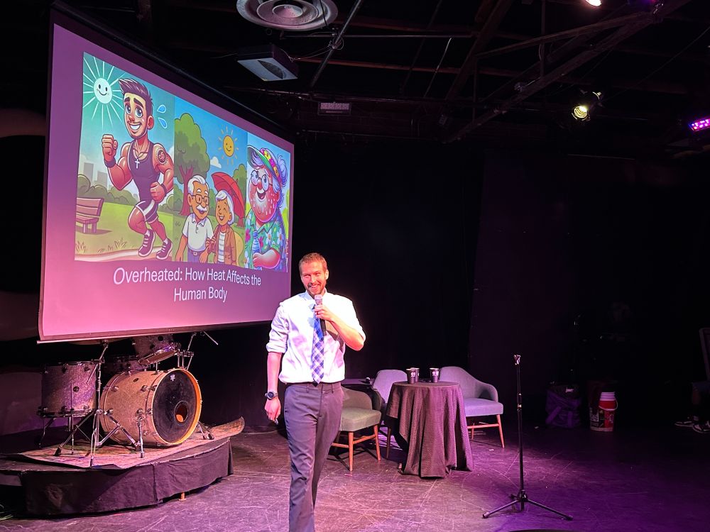 A speaker is smiling while presenting in front of a screen with images of people and the headline, "Overheated: How Heat Affects the Human Body." 
