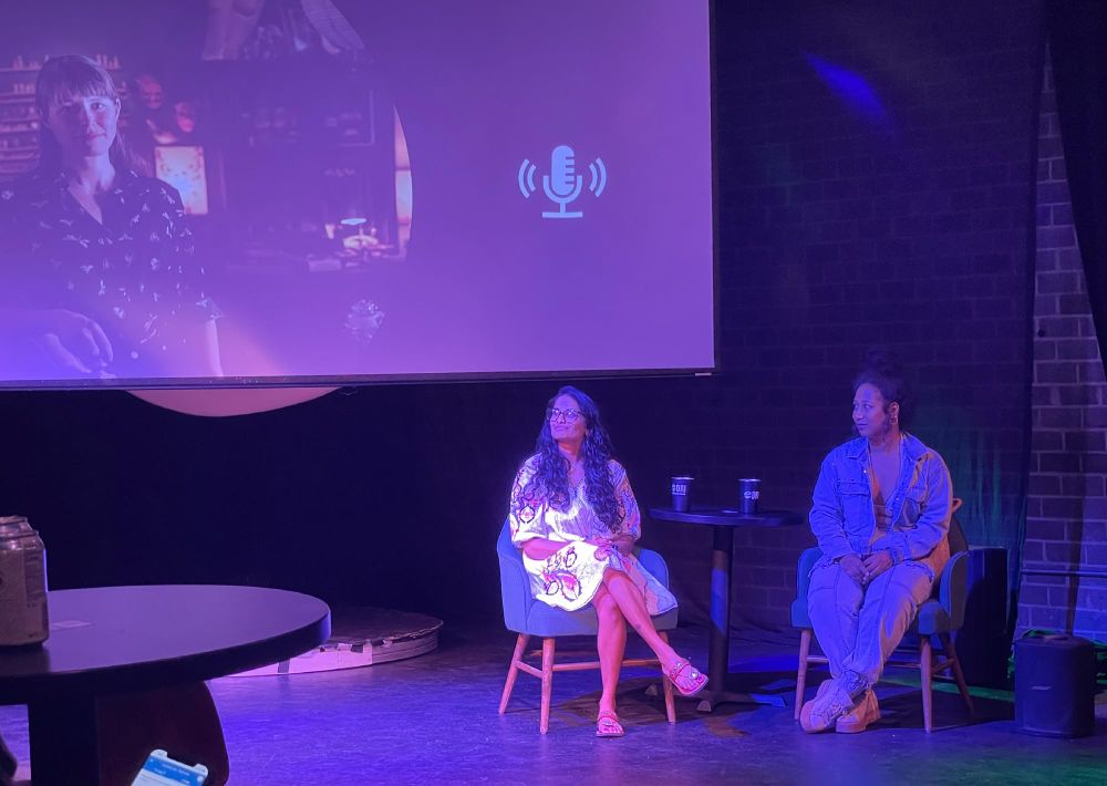 Tiffany and CoCo seated on stage at a during Q&A at the event, with a projected image of Jessica who joined virtually on the screen behind them.