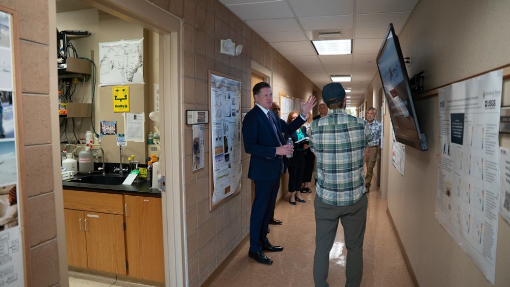 NSHE Chancellor McNair engaging with researchers in a hallway surrounded by scientific posters and equipment can be seen through an open doorway.