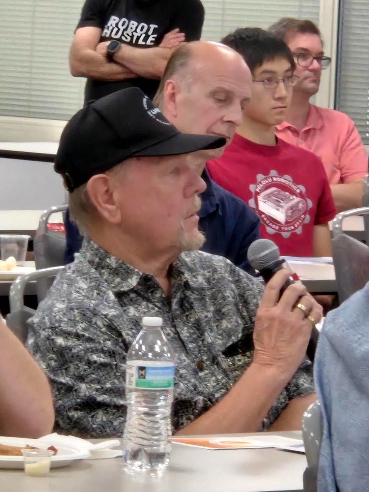 Man wearing a cap, holding a microphone  with three others seated near him.