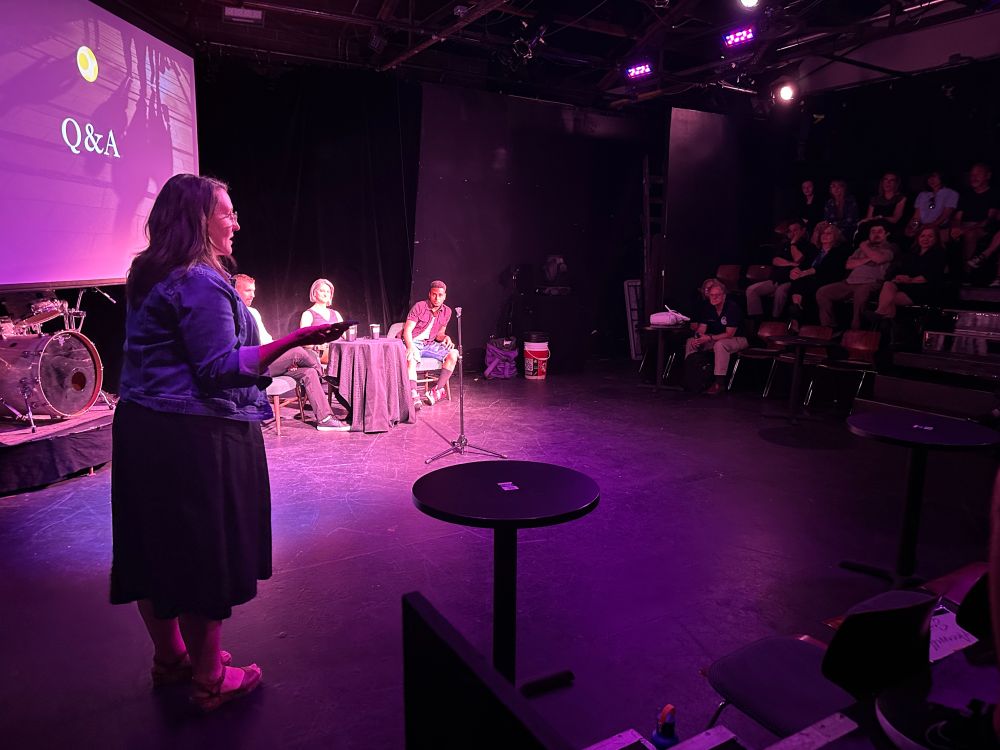 The event host is addressing the audience in front of a table with three speakers and the text Q&A on the screen behind them. 