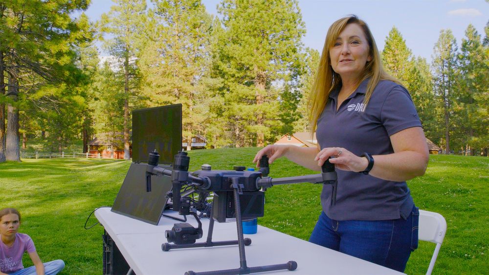 Scientists Teresa stands next to a table holding a drone. Trees and green grass behind her.
