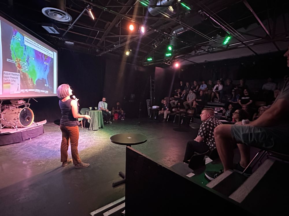 A speaker is on stage in front of a presentation on a screen with the audience looking on. 