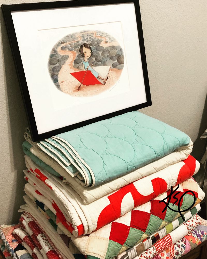 A framed art print by Sophie Blackall. A dark haired girl paddles an open book, down a make believe stream. The art piece sits on a stack of colorful quilts.