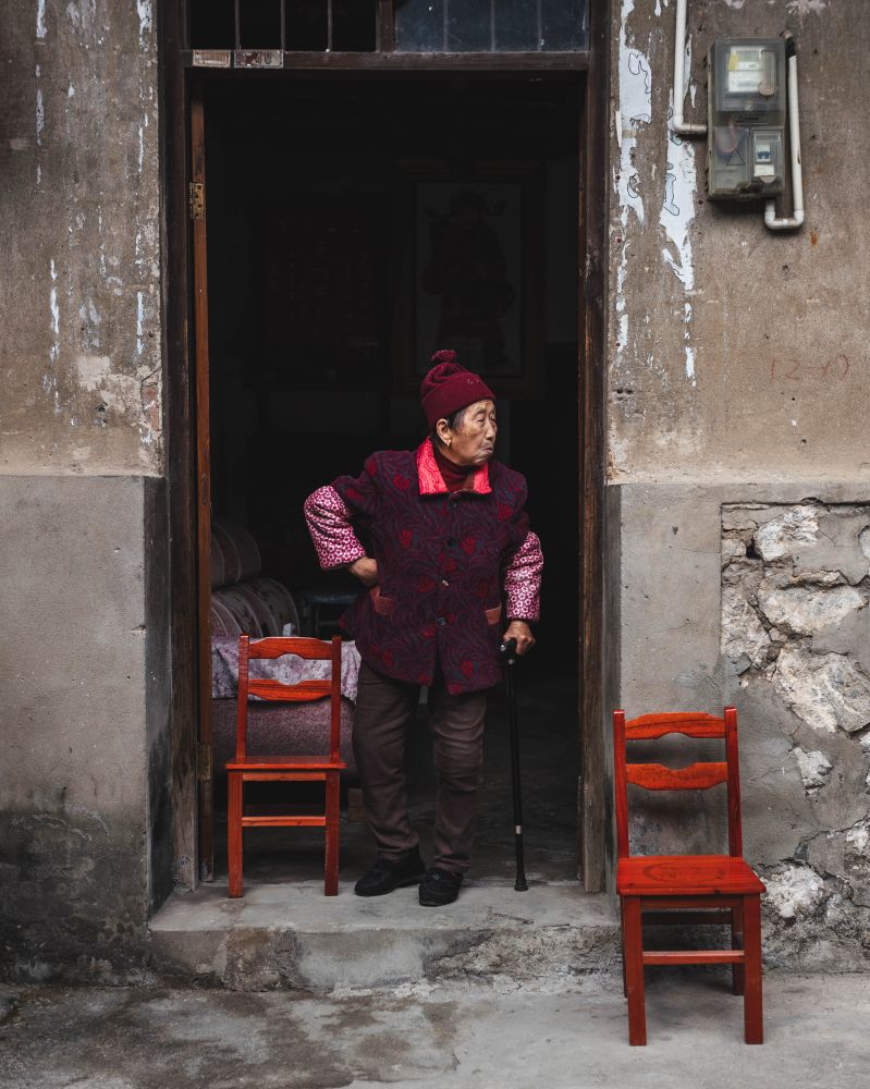 Elderly woman from Guilin, China, in red clothing. Her right arm rests on her hip and she’s leaning on her black cane. She is standing at her open doorway with two red chairs, one each at her side.