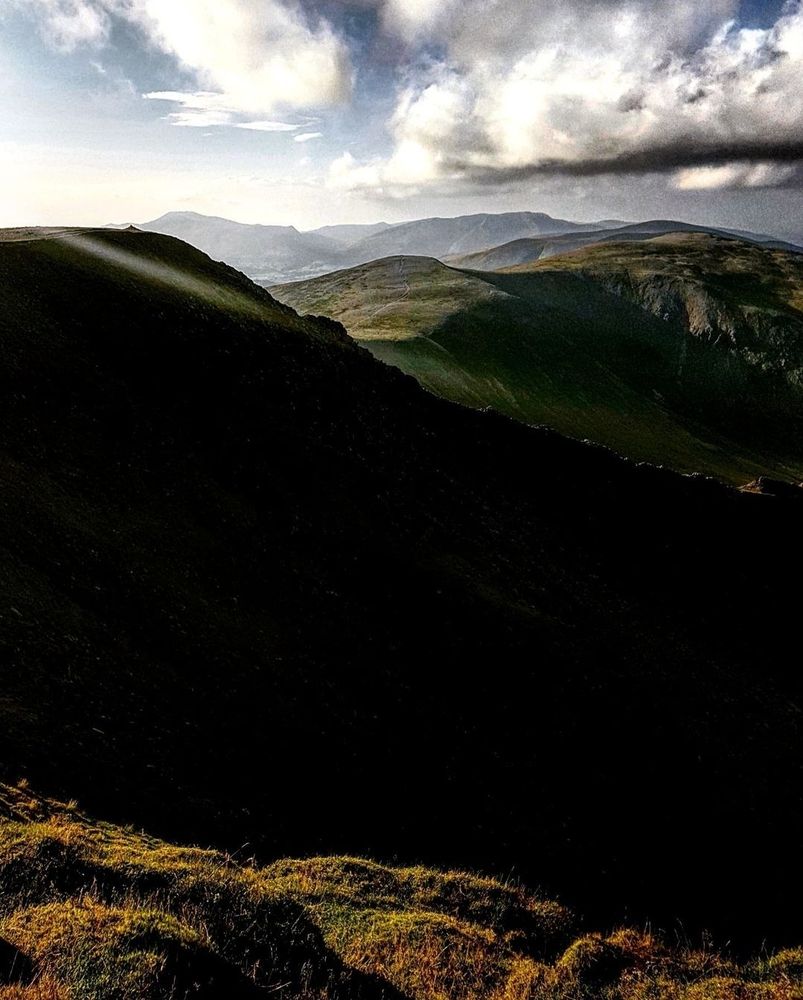 A mountainscape. In the foreground, there is a sliver of fellside, lit up golden by the sun. Behind it, the mountain rises, pure black in silhouette, the lit fells stretching out behind.