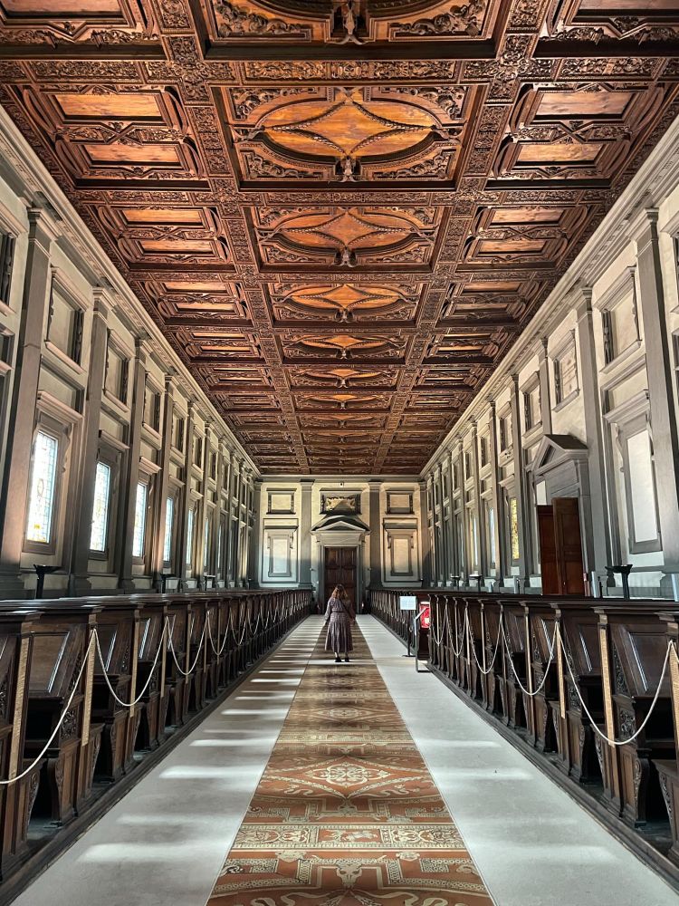 A large long room. On the long sides of the room are dark wooded desks and seats which are roped off. The floor is carpet which is cut to reveal a mosaic strip down the middle. There are large windows with stained glass in them down the whole stretch of the room. The ceiling is carved wood. Kate stands in the middle as a reference for size. She looks very small in comparison to the room. 