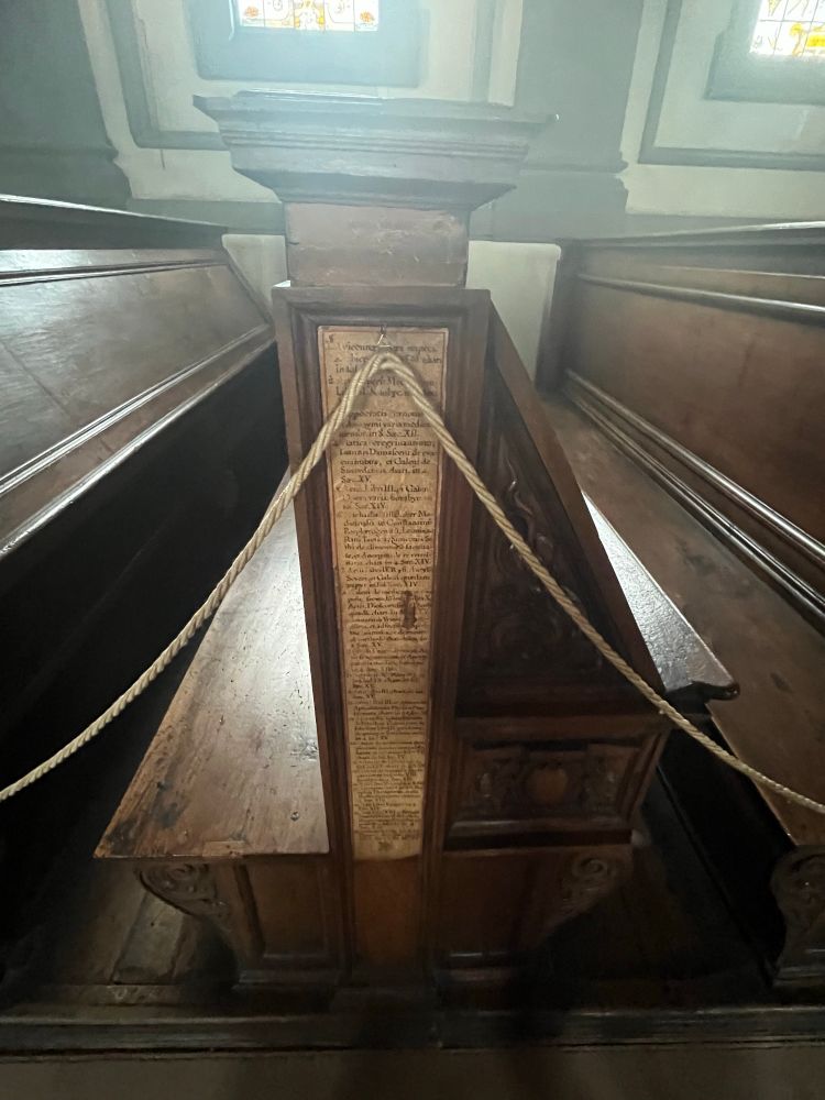 A close up on the tilted desks and seats. The desk is joined to its corresponding seats like a carrell. The seat is made up of a communal bench with a high back. The high back of the bench is flush with the desk behind it. They are a dark wood with carvings. On the side of the each bench’s back rest is a hand writing book list detailing which books are fixed to the desks.