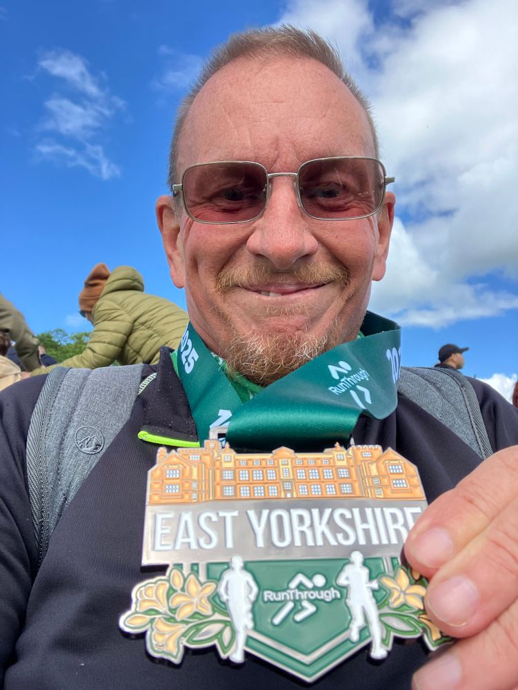 Post race selfie showing a slightly tireder but very contended grin proudly showing off my lovely finishers medal, a lovely bronze coloured design of The Hall over the words East Yorkshire white on black above a design of two runners running on the greensward. 