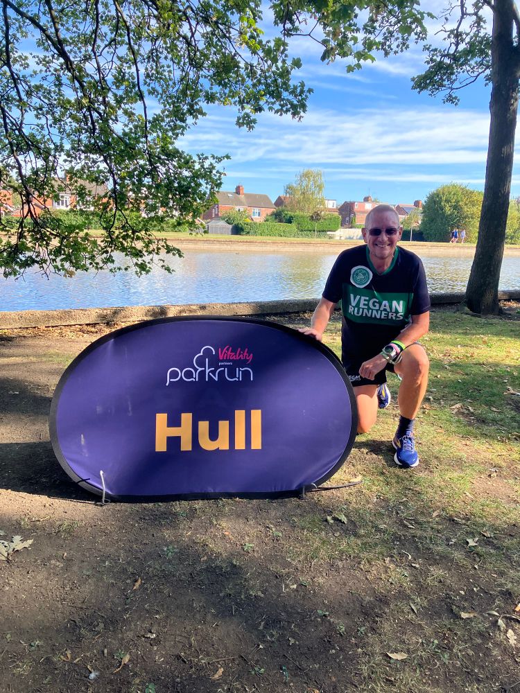 Mark in his Vegan Runners kit with Green Runners logo beaming happily next to the Hull parkrun flag, with gorgeous blue sky and the East Park lake behind him