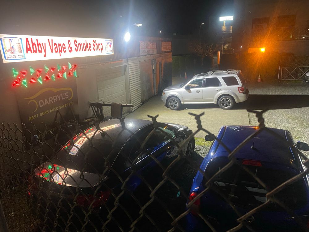 a “car sales lot” brightly illuminated to near-zero effect, additional signage present for a vape store and smoke shop that closes before sundown