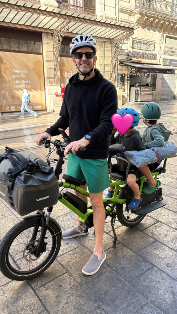 Man on a green cargo bike with a big bag in front and two kids on the back with a beach umbrella