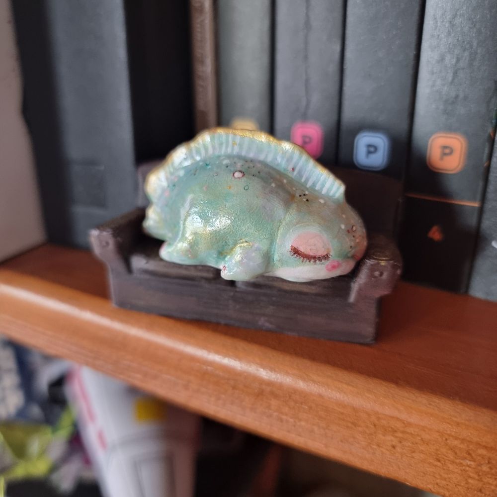 A small green painted amphibian-like figurine appears to be sleeping on a tiny beat-up brown two-seater couch. There are books behind the couch, and a wooden shelf below