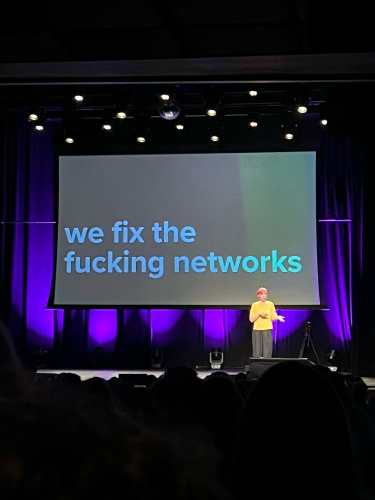 Writer/researcher Erin Kissane wearing. Bellow shirt with coral pink hair standing on stage at XOXO Fest in Portland. The stage has purple velvet curtains. On the screen behind Erin is says “we fix the fucking networks”.