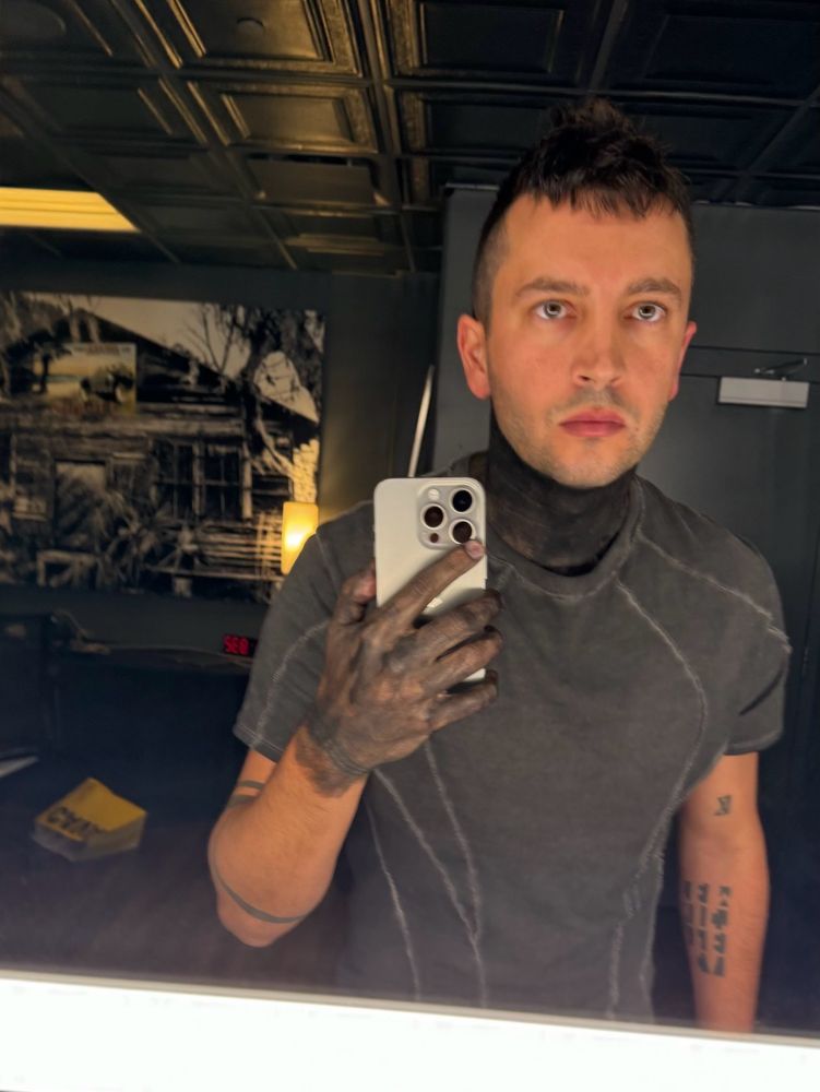 tyler joseph dressed in a black tshirt with his black paint on his hands and neck. he is taking a mirror selfie. his expression is blank. 