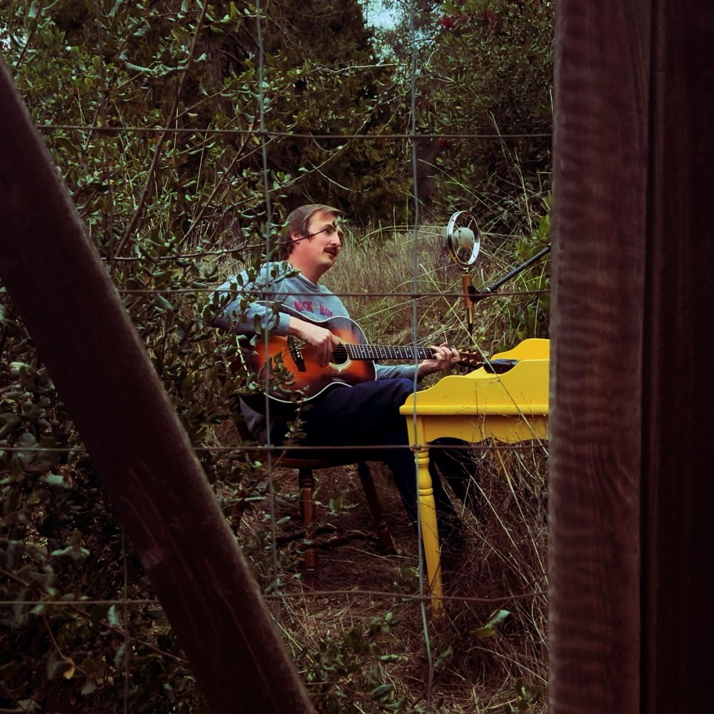 Grover sitting outdoors, playing guitar at a desk and singing into an Ear Trumpet Microphone. Viewed through a wood and chicken wire fence.