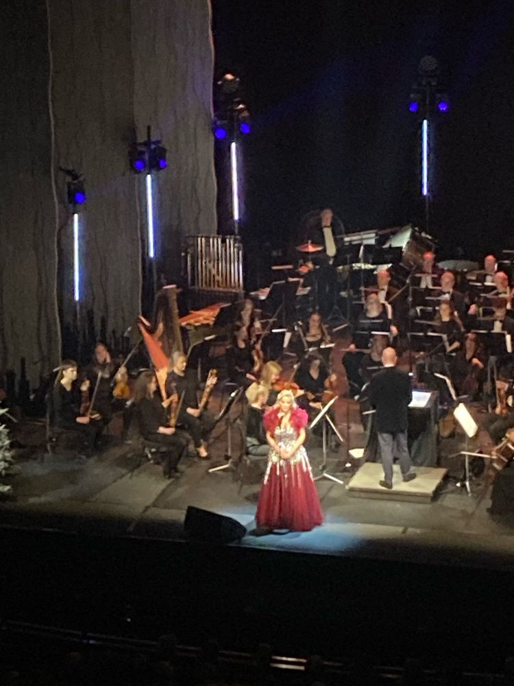 Photo shows an orchestra onstage with singer Celine Byrne at the microphone, in a red and silver dress.
