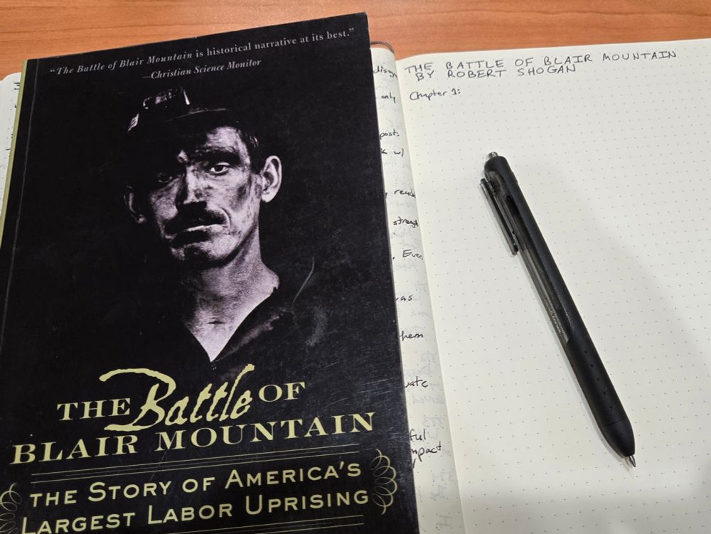 The book "The Battle of Blair Mountain" by Robert Shogan next to an empty page of a notebook with the book's title at the top. A pen sits on the notebook.
