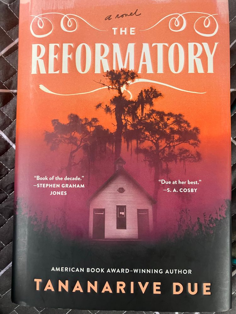 Cover of The Reformatory by Tananarive Due. White house with two doors pictured in front of moss dripping trees on a penumbra of orange to purple to charred black base. 