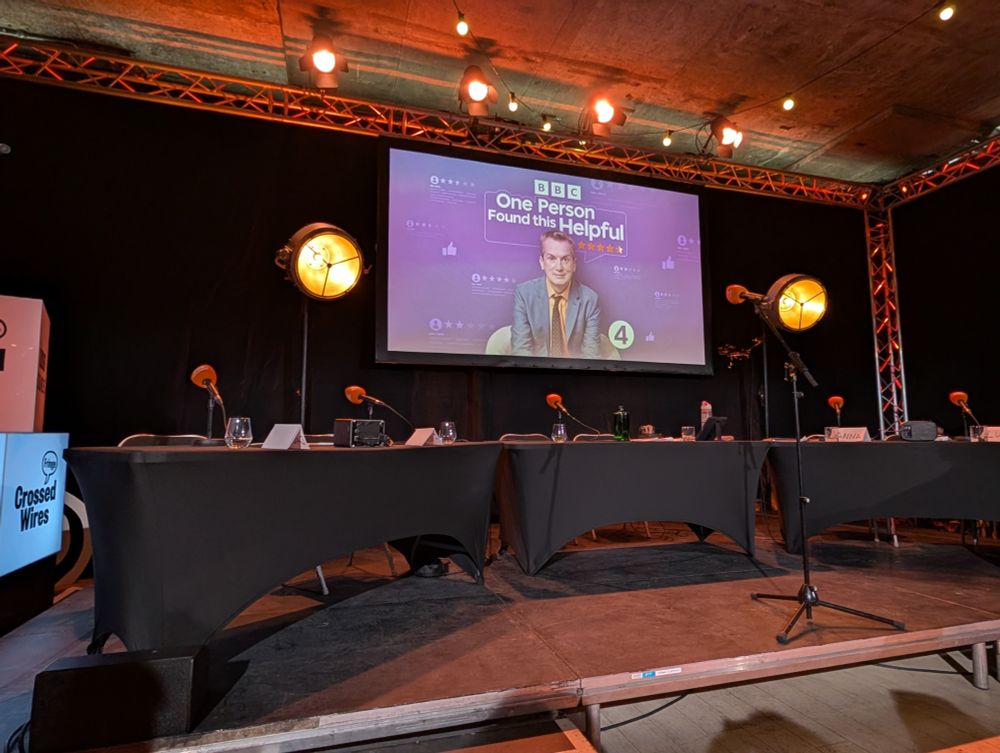 Stage setup for 'One Person Found This Helpful'. Three black clothed tables with microphones are lined up with a projected image of Frank Skinner and the show's title behind them.