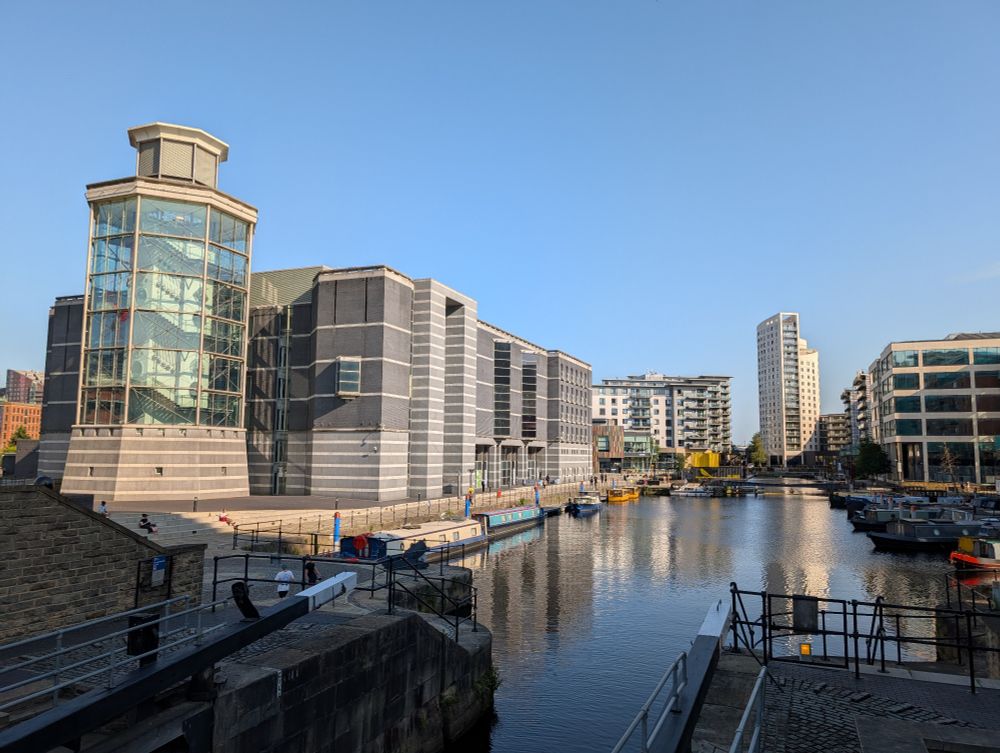 Leeds Dock, a large water basin surrounded by modern buildings. The sky is blue and cloudless. The buildings are reflected in the calm water. 