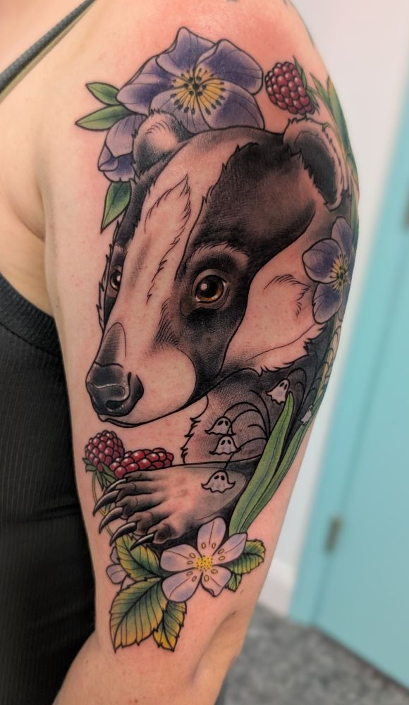 A tattoo on an upper arm of a Eurasian badger eating raspberries, with a few hellebore and lily of the valley surrounding.