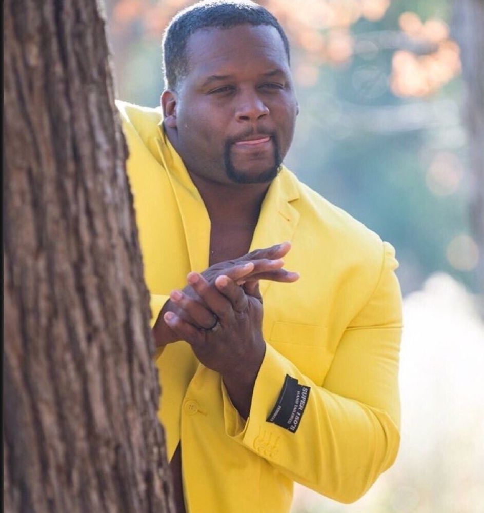 Guy in yellow suit behind tree rubbing hands together meme