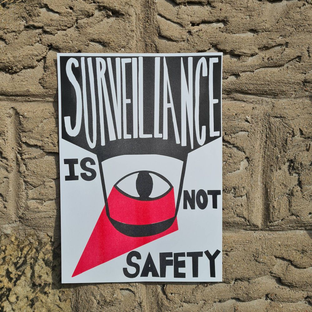 In the centre of the poster is the black silhouette of CCTV camera embedded into the ceiling, with a black eye in the centre. A red triangle stretches out from the eye into the bottom left corner. White text cut out of the ceiling above the camera reads: Surveillance and beneath this large block text in black reads ‘Is Not Safety’.
