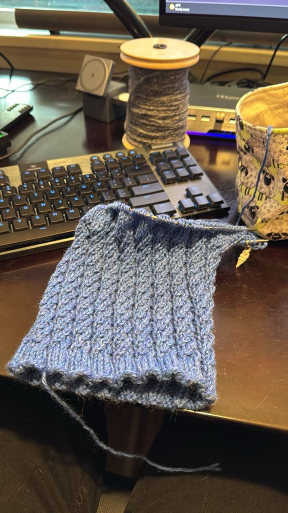 Denim blue merino two horned elfish hat, unfinished, on the needle, resting on a computer desk with misc. fiber arts stuff strewn about.