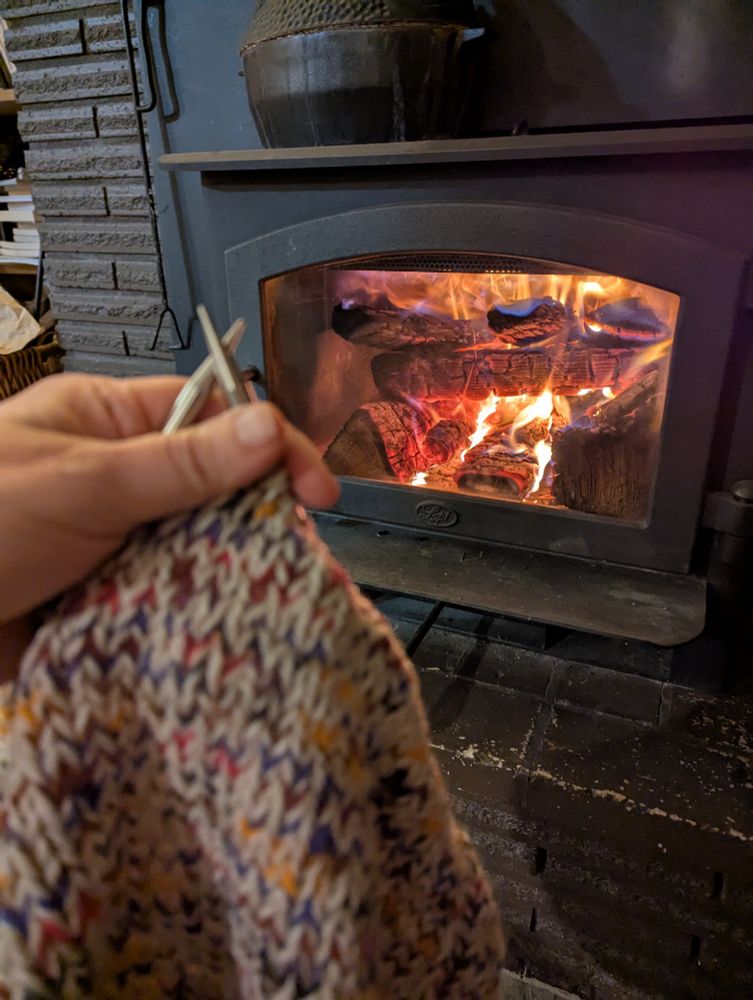 White and multi colored knitting held up in front of a cozy wood stove with a fire burning. 