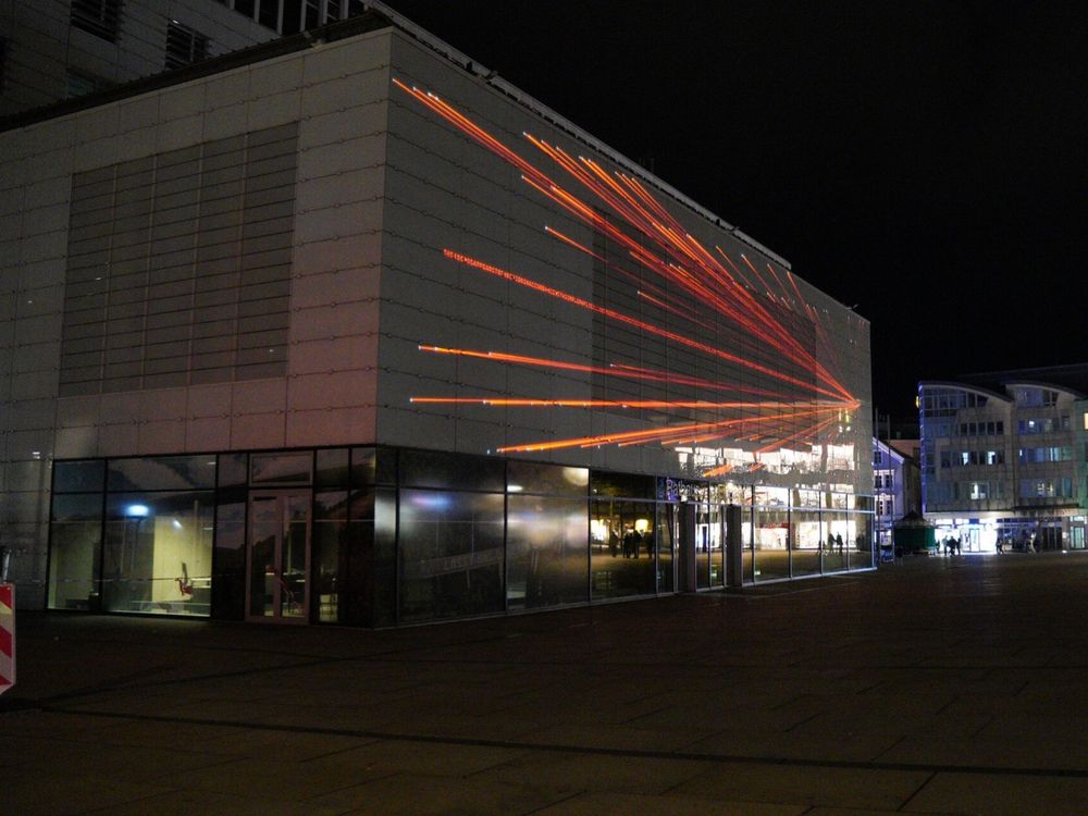 Das Foto wurde am Abend oder nachts gemacht, da der Himmel schwarz ist. Man sied eun quaderförmiges, helles Gebäude, auf das mit rotem Licht Striche projiziert werden. Im unteren Teil der Fassade spiegeln sich erleuchtete Fenster. 