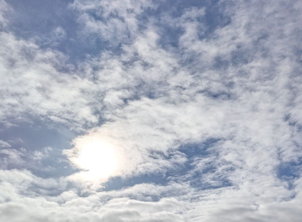 Blauer Himmel mit vielen Wolken. Hinter einer ist die Sonne versteckt. Man kann sie erkennen, da diese Wolke leuchtet. 