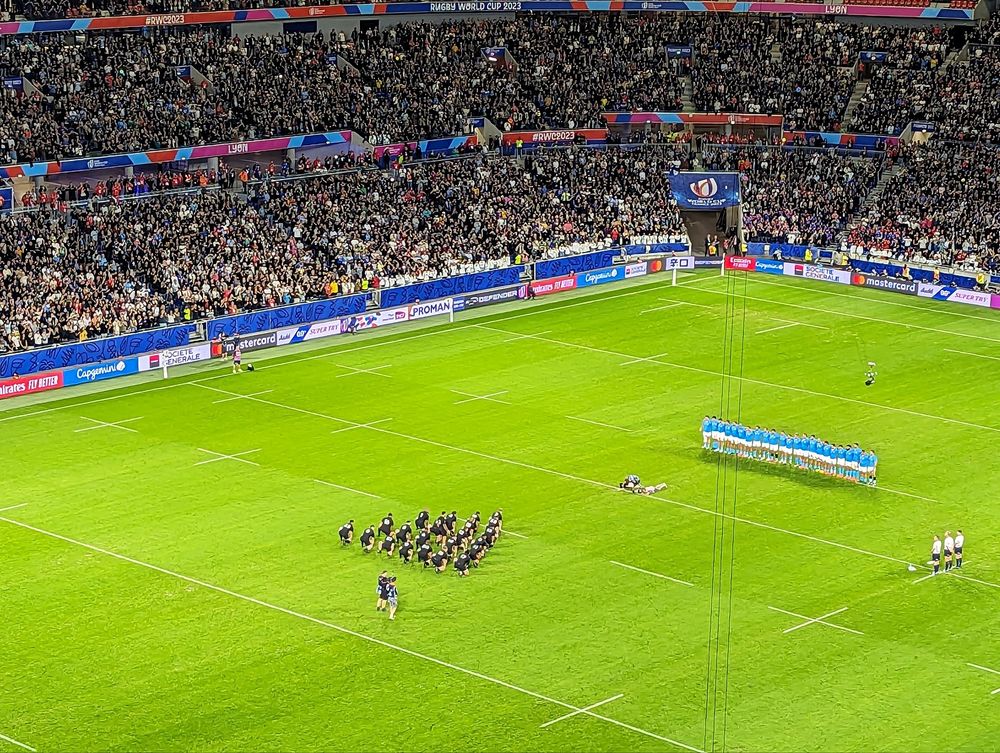 Ein vollbesetztes Stadion. Auf der Rasenfläche steht eine blauweiß gekleidete Mannschaft in einer Reihe. Ihr gegeüber steht eine schwarz gekleidete Mannschft in der Formation einer Pfeilspitze. 3 Männer stehen quer zu den Mannschaften. 