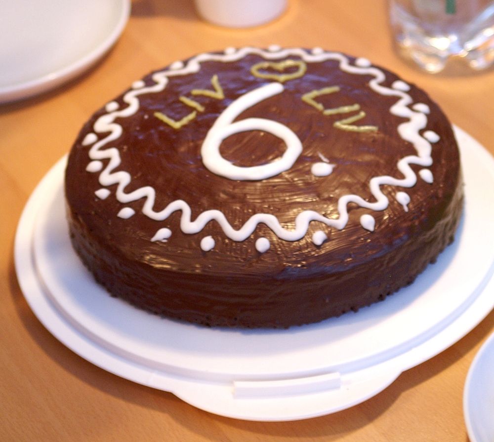 Auf eunem weißen Torteteller liegt ein mit Schokolade überzogener Kuchen. Er ist mit Zuckerguss verziert. In der Mitte steht eine 6, darüber 2x der Name Liv und ein Herz. 