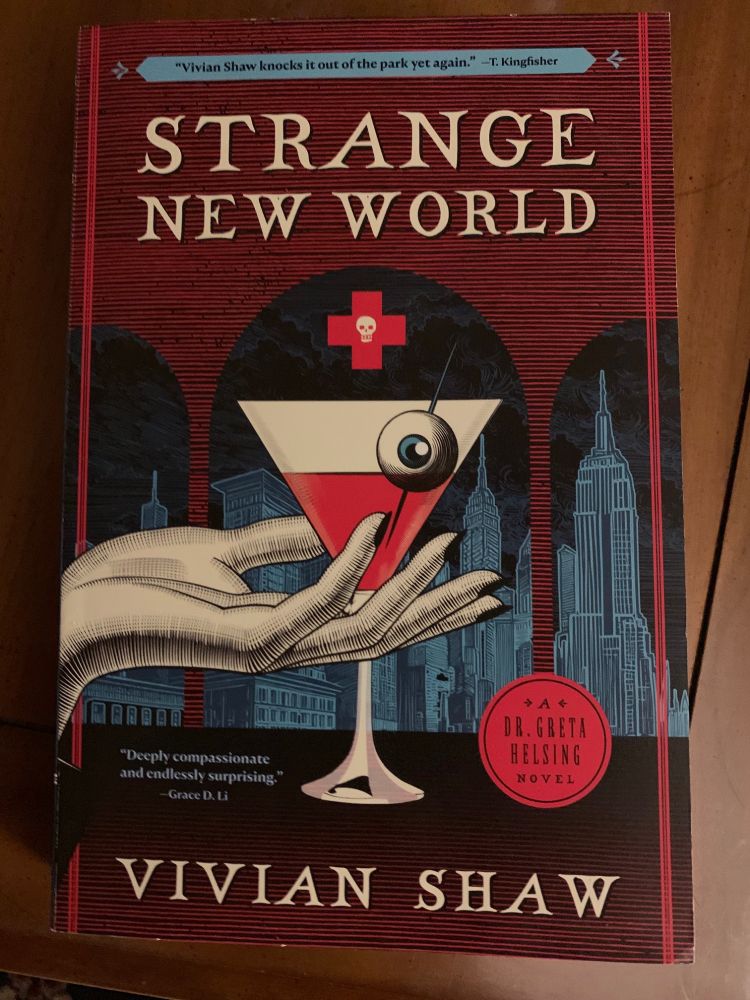 Strange New World, by Vivian Shaw. The cover is in a colored woodcut-style. A human hand holds a wineglass that contains a red liquid with an eyeball garnish, underneath a red cross with a skull. Skyscrapers loom in the background.