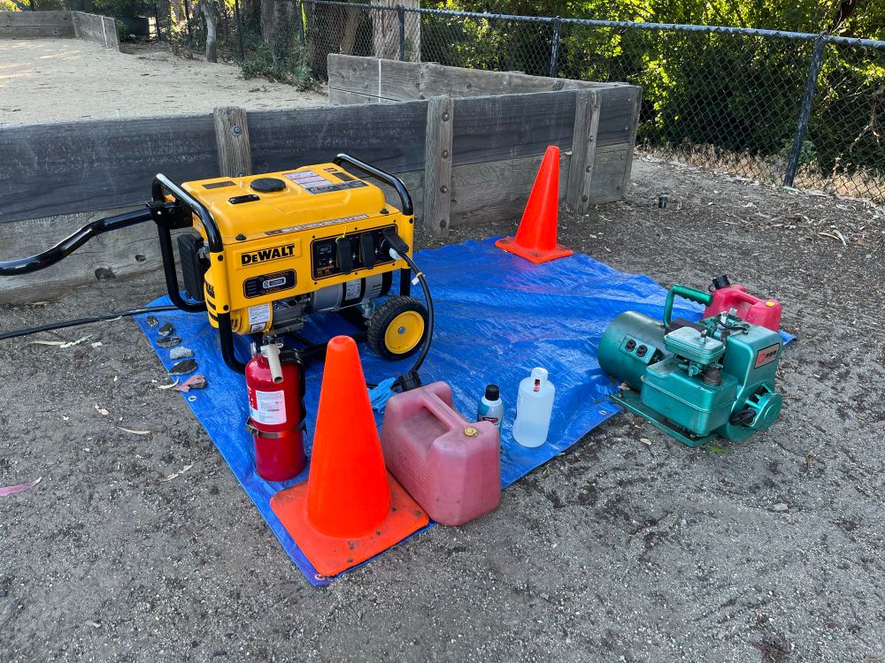 A gas-powered generator on a blue tarp. A fire extinguisher and a can of fuel are placed next to it. Two traffic cones highlight the area. A smaller, older-looking generator is off to the site, but isn’t connected. 