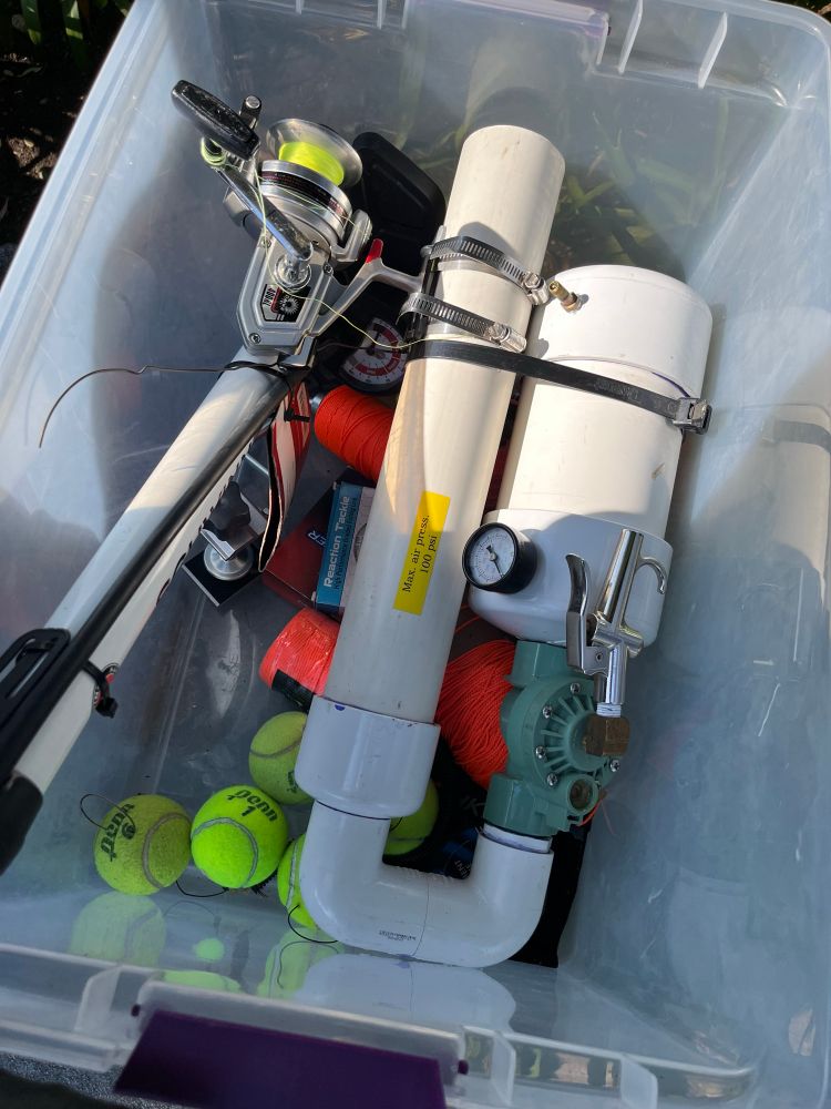 Storage box containing an air pump, several yellow tennis balls, and a large device made of large white PVC tubing. A valve handle, pressure gauge, and a fishing rod reel are attached to the large device. 