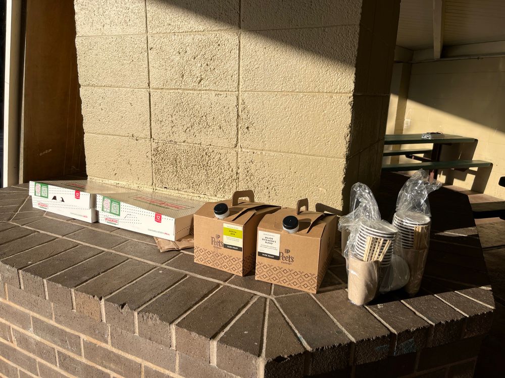 Two boxes of donuts and two containers of coffee with cups and stirrers sitting on a flat concrete wall. 