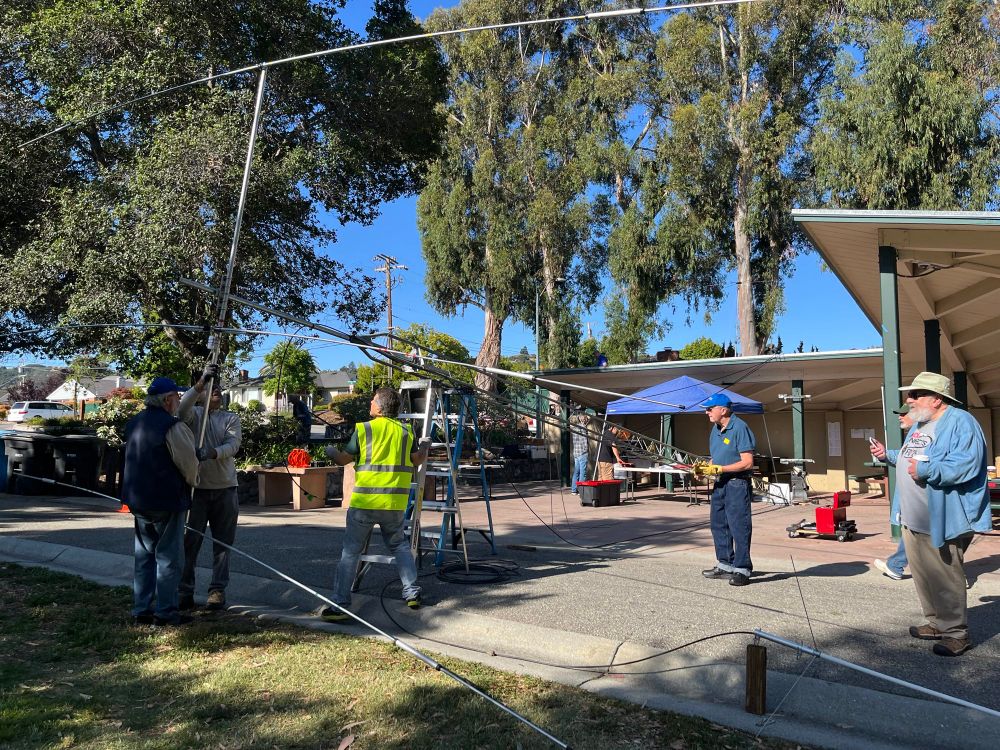 The people are handling a large Yahi antenna that they mount onto a rigging tower that is leaning on a ladder at a ca. 15 degree angle. In the background is the park shelter where the radio stations are being set up. 
